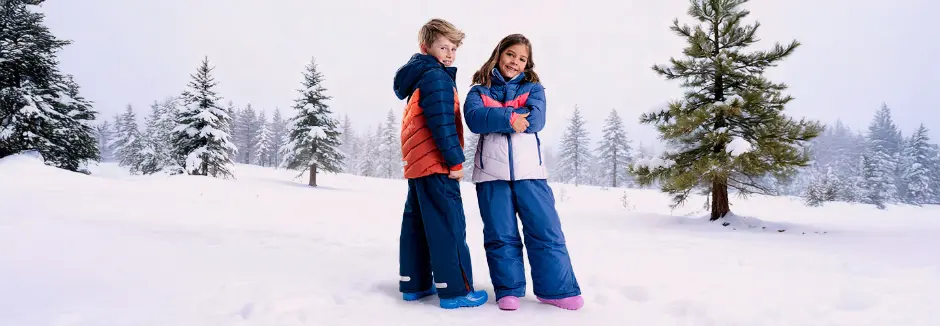 Zwei Kinder in bunten Winterkleidern stehen in einer verschneiten Landschaft, umgeben von hohen immergrünen Bäumen und einer nebligen Atmosphäre.